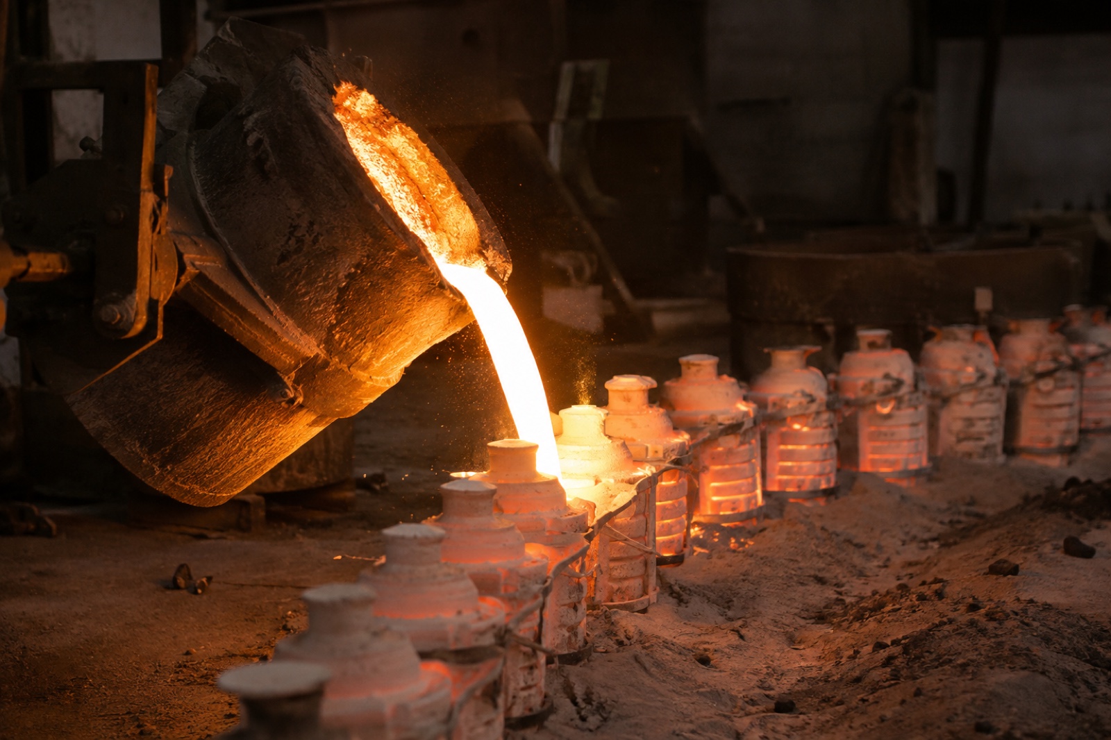 Molten steel poured from a ladle into a row of preheated ceramic investment casting shells on a foundry floor, the shells glowing from residual heat